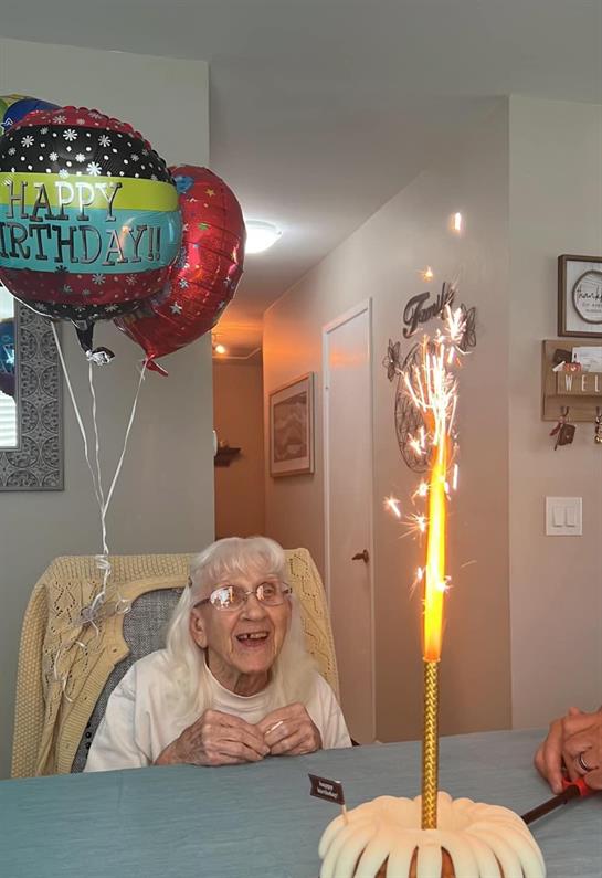 An elderly woman joyfully celebrates her birthday with balloons and a sparkling candle.