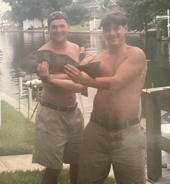 Two shirtless men smile with a large fish by the canal, celebrating their catch.