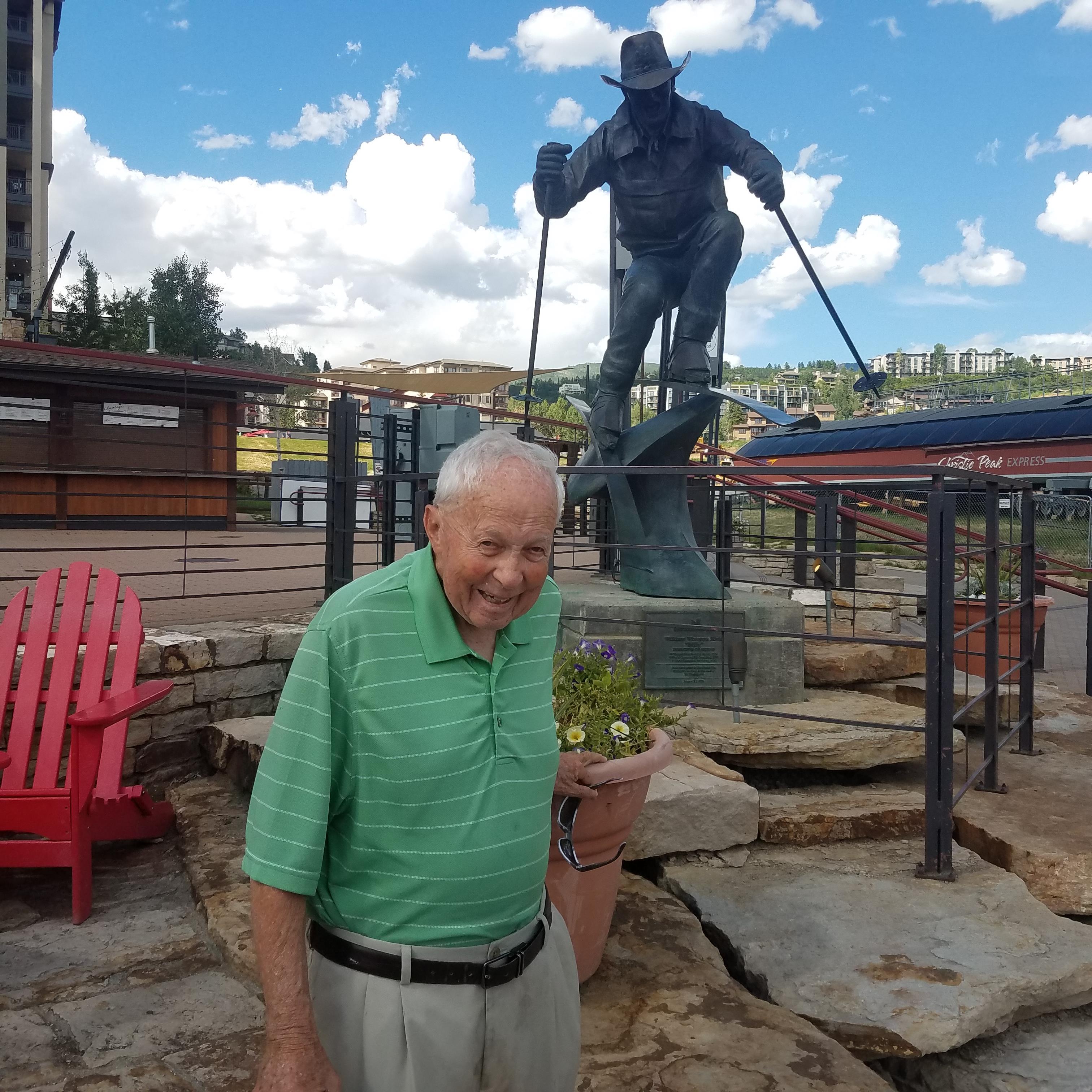 Joyful elderly man stands in front of a statue while holding a small plant in a sunny area.
