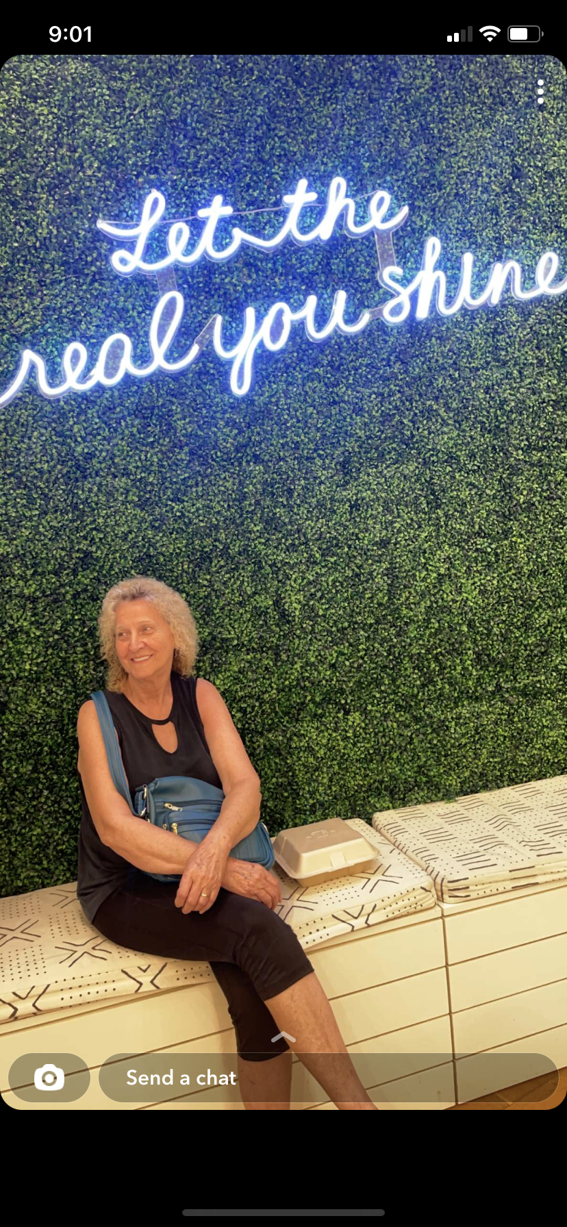 An elderly woman with curly hair relaxes on a bench against a greenery wall with motivational text.