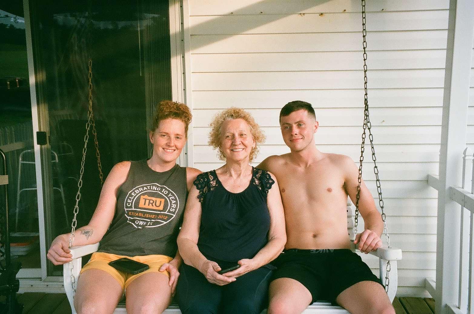 Three family members are seated together on a porch swing, sharing smiles and laughter.