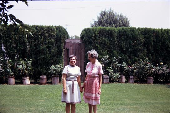 Two women stand in a green garden, enjoying a sunny day while holding hands and smiling.