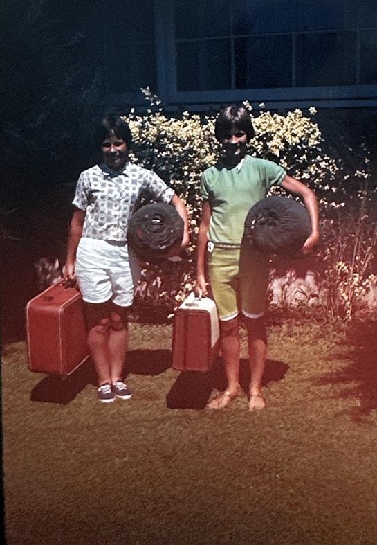 Two kids stand with suitcases and rolled sleeping bags, ready for adventure in their yard.
