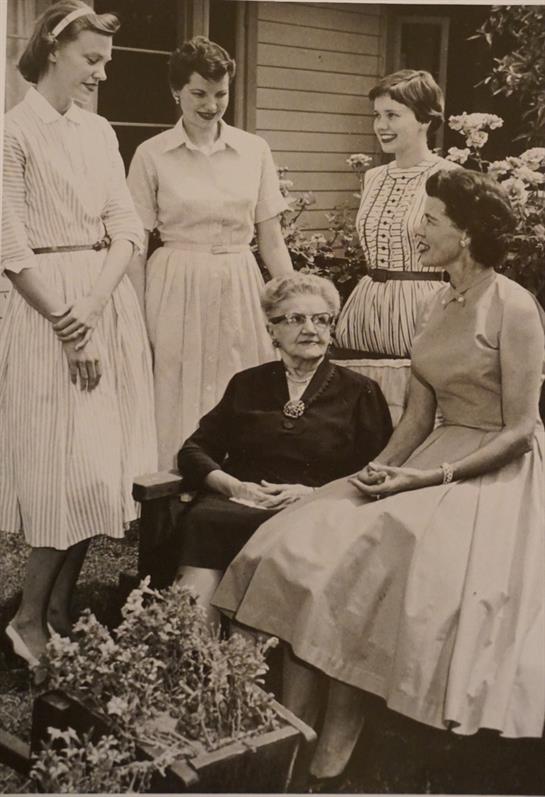 Five women in vintage dresses engage in conversation with an elder woman in a garden setting.