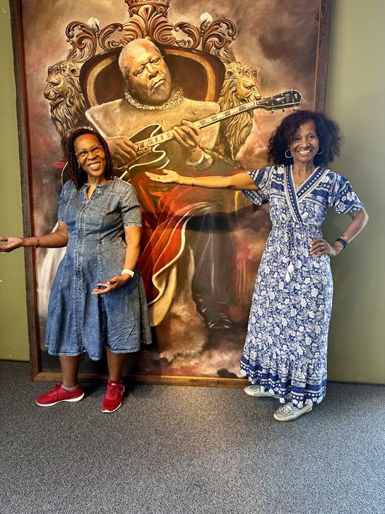 Two women stand with cheerful expressions beside an artistic mural of a guitarist.