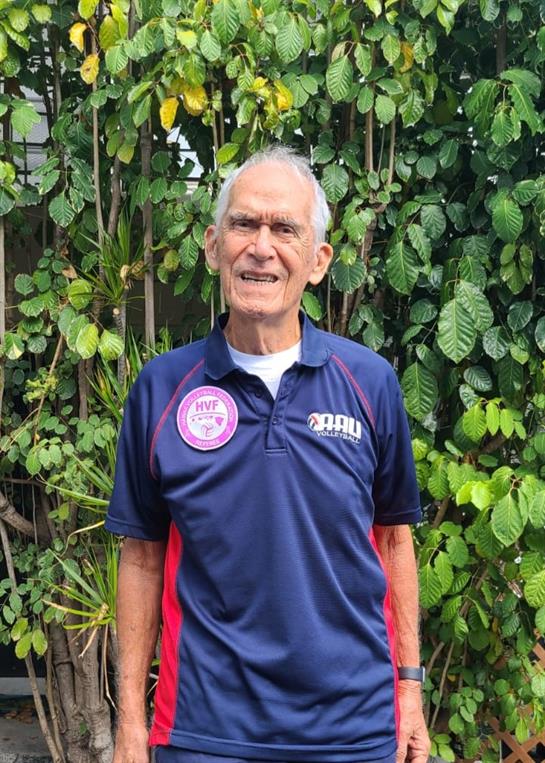 Elderly man stands happily in a blue sports shirt amidst vibrant green leaves.