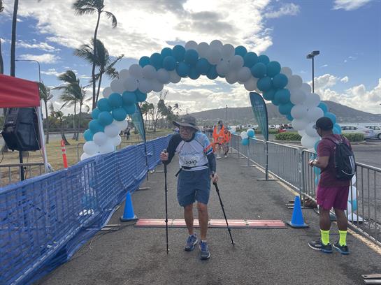 A person using walking poles arrives at the finish line with a colorful balloon arch overhead.