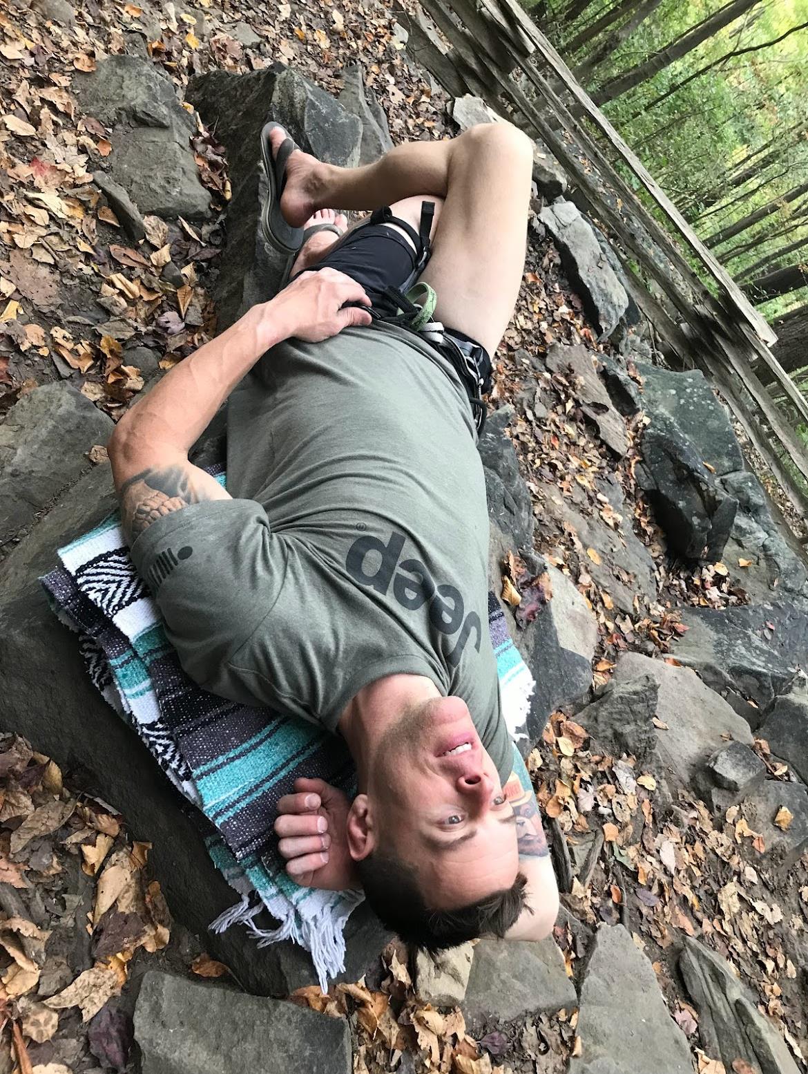 A person lies on a blanket, resting on large rocks surrounded by trees and leaves.