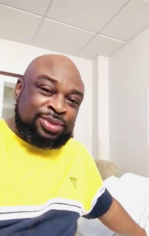 A man relaxes indoors, smiling brightly in his vibrant yellow shirt.