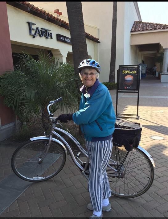 An elderly person in a blue jacket and helmet joyfully stands beside a bicycle outside shops.