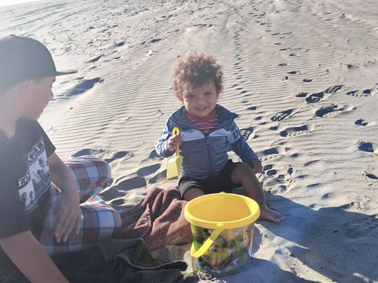 A young child sits on the sand with a yellow bucket, enjoying a sunny day at the beach.
