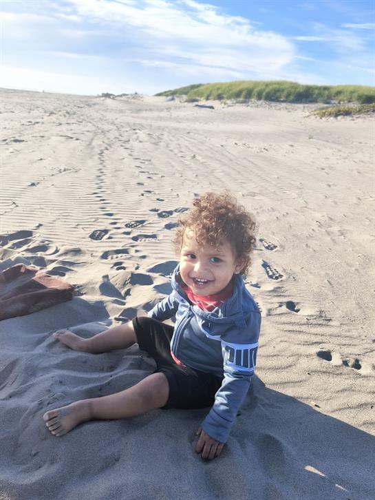 A happy child sits on the sandy beach, playing in the sand under a clear blue sky.