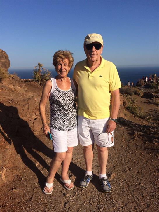Older couple poses happily at a rocky lookout with a beautiful ocean backdrop on a clear day.