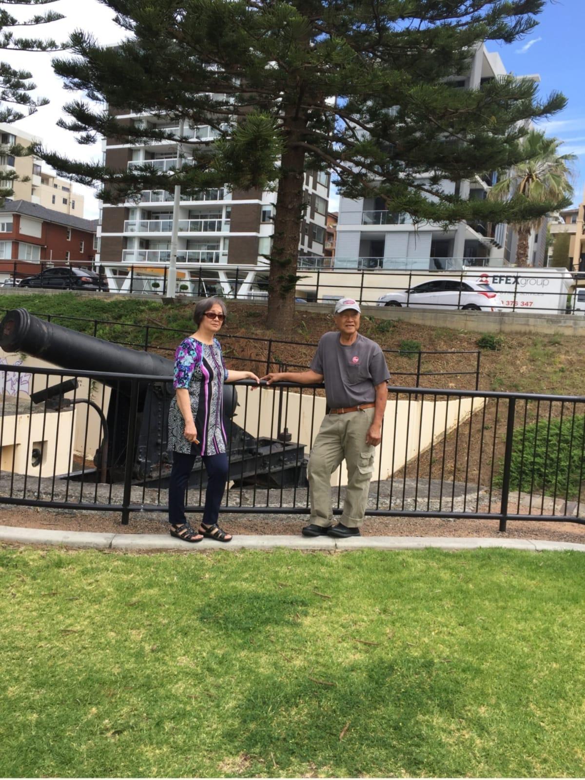 Two individuals stand next to a historic cannon in a park surrounded by modern buildings.