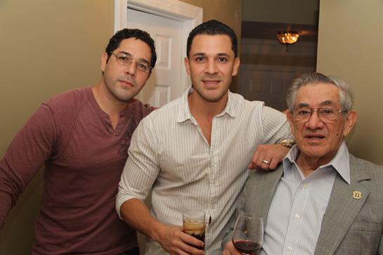 Three men stand together holding drinks, sharing laughter and conversation in a cozy setting.