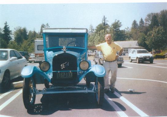 A man in a yellow shirt stands proudly next to a classic blue car in a parking area.