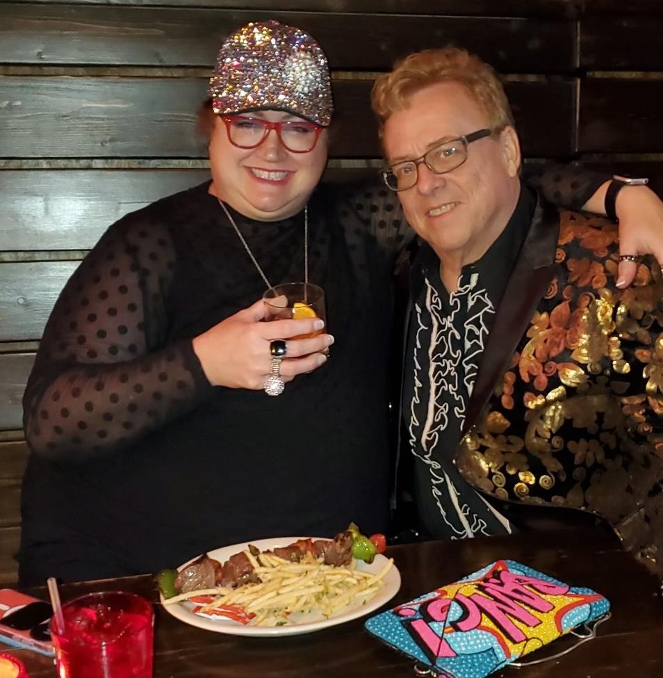 Two friends smile while enjoying dinner together at a vibrant eatery, sharing drinks and food.