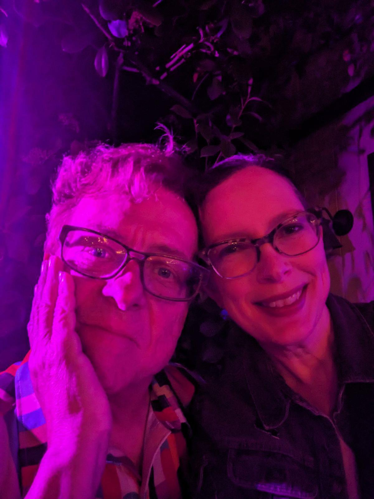 Two friends pose under colorful lights at a lively evening event.