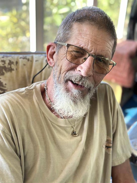 A cheerful elderly man in glasses and a light shirt smiles while sitting comfortably.