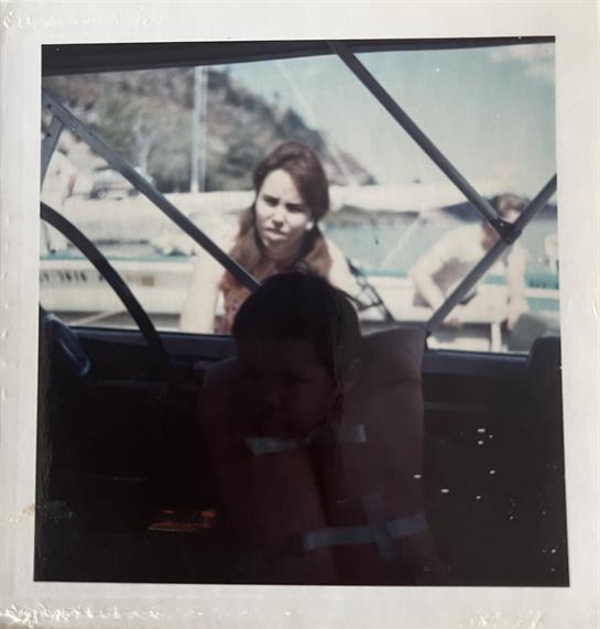 A woman leans over a boat, looking curiously at a man seated inside, enjoying a sunny day.