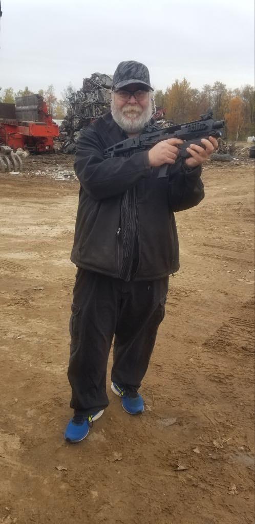 Individual stands in a junkyard, gripping a firearm and wearing dark clothing.
