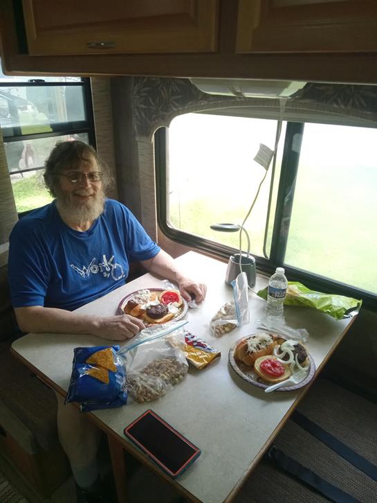 A man sits at a table in an RV, enjoying a meal with fresh ingredients and snacks.