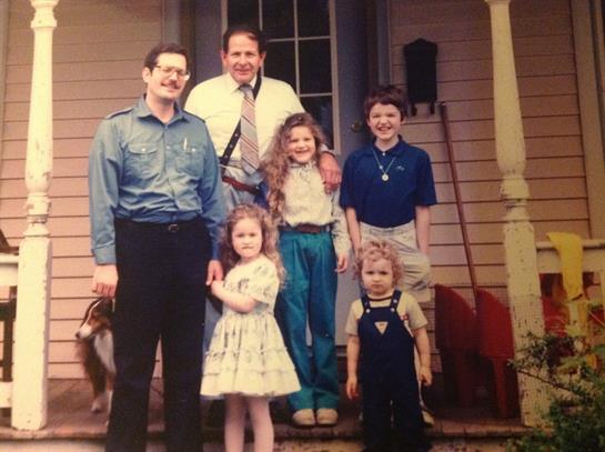 A group of five adults and children pose together on a porch during a sunny afternoon.