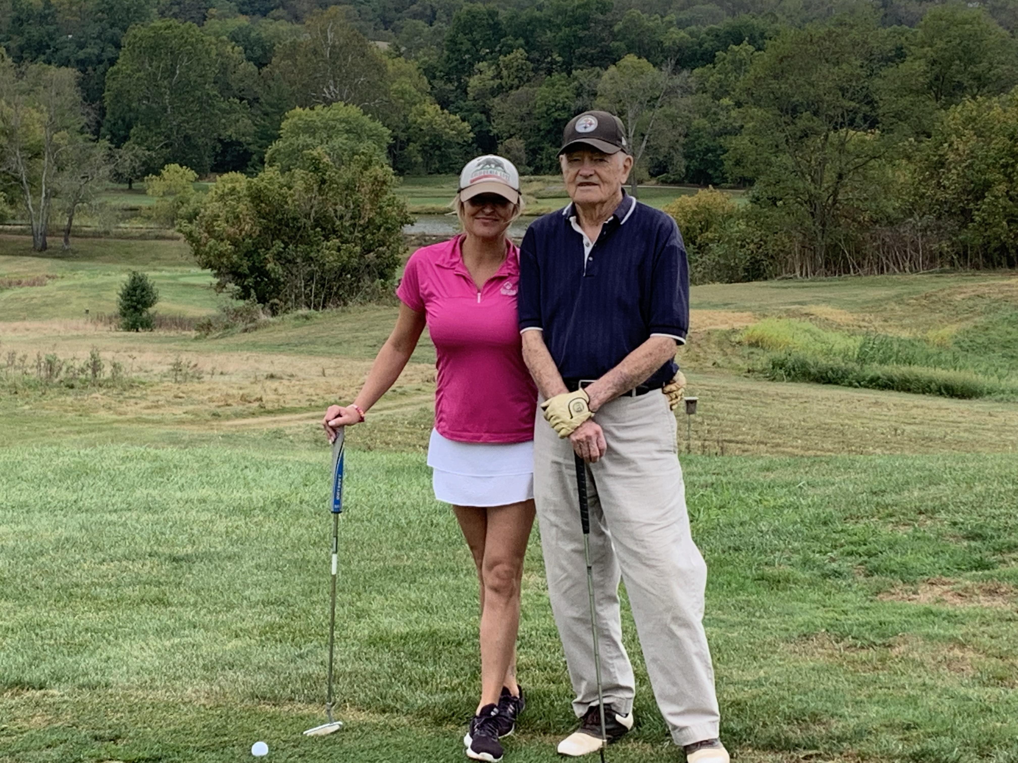 A man and woman pose with golf clubs, smiling on a sunny day surrounded by greenery.