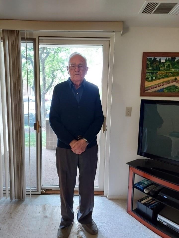 An elderly man with glasses stands silently in a well-lit living room by a sliding door.