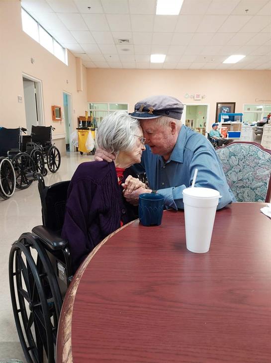 In a care facility, an elderly couple affectionately touches foreheads, showing their bond.