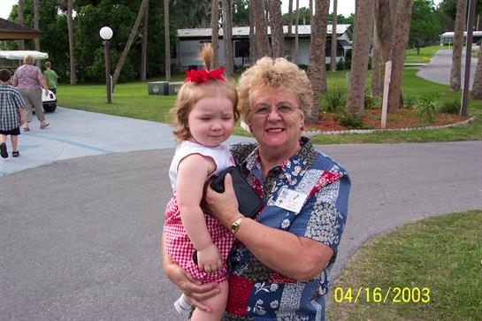 A joyful elderly woman holds a young girl in a park, surrounded by green grass and trees.