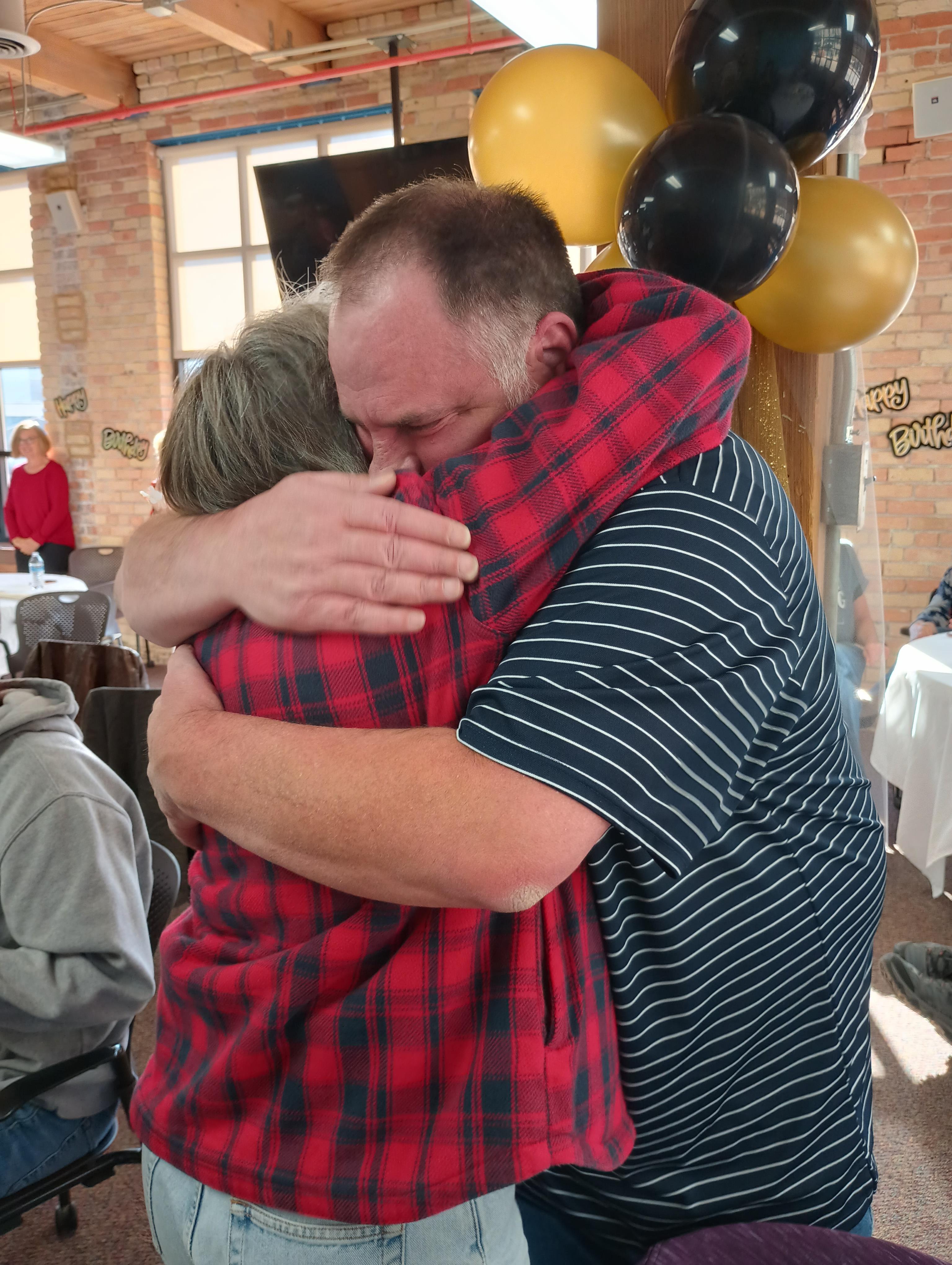 Two people embrace each other warmly, sharing a heartfelt moment in a lively indoor setting.