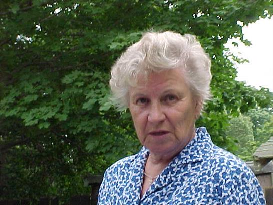 A senior woman with curly gray hair wears a blue patterned shirt while standing outside.