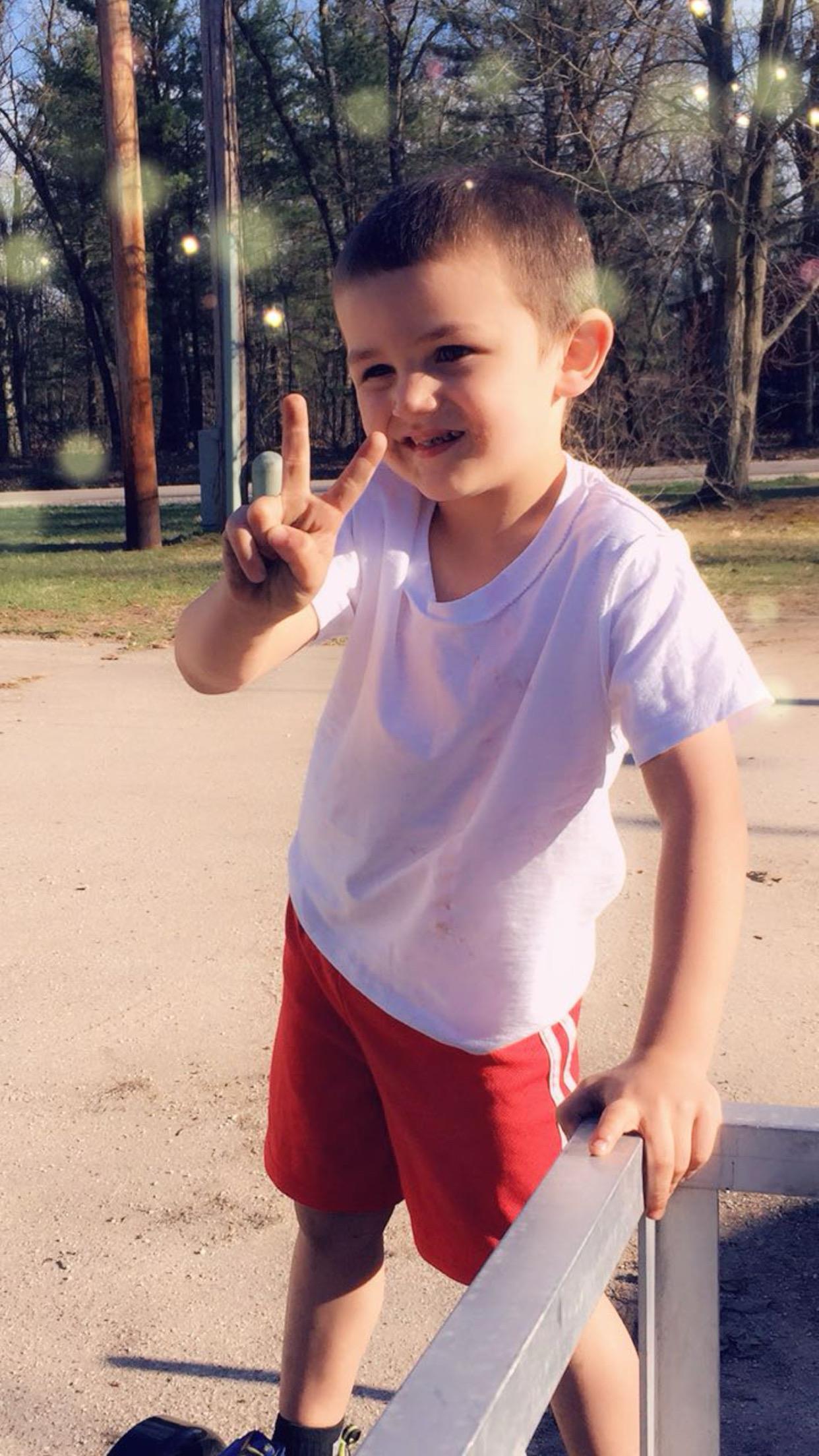 A cheerful child stands at a park, smiling and making a peace sign with his fingers.