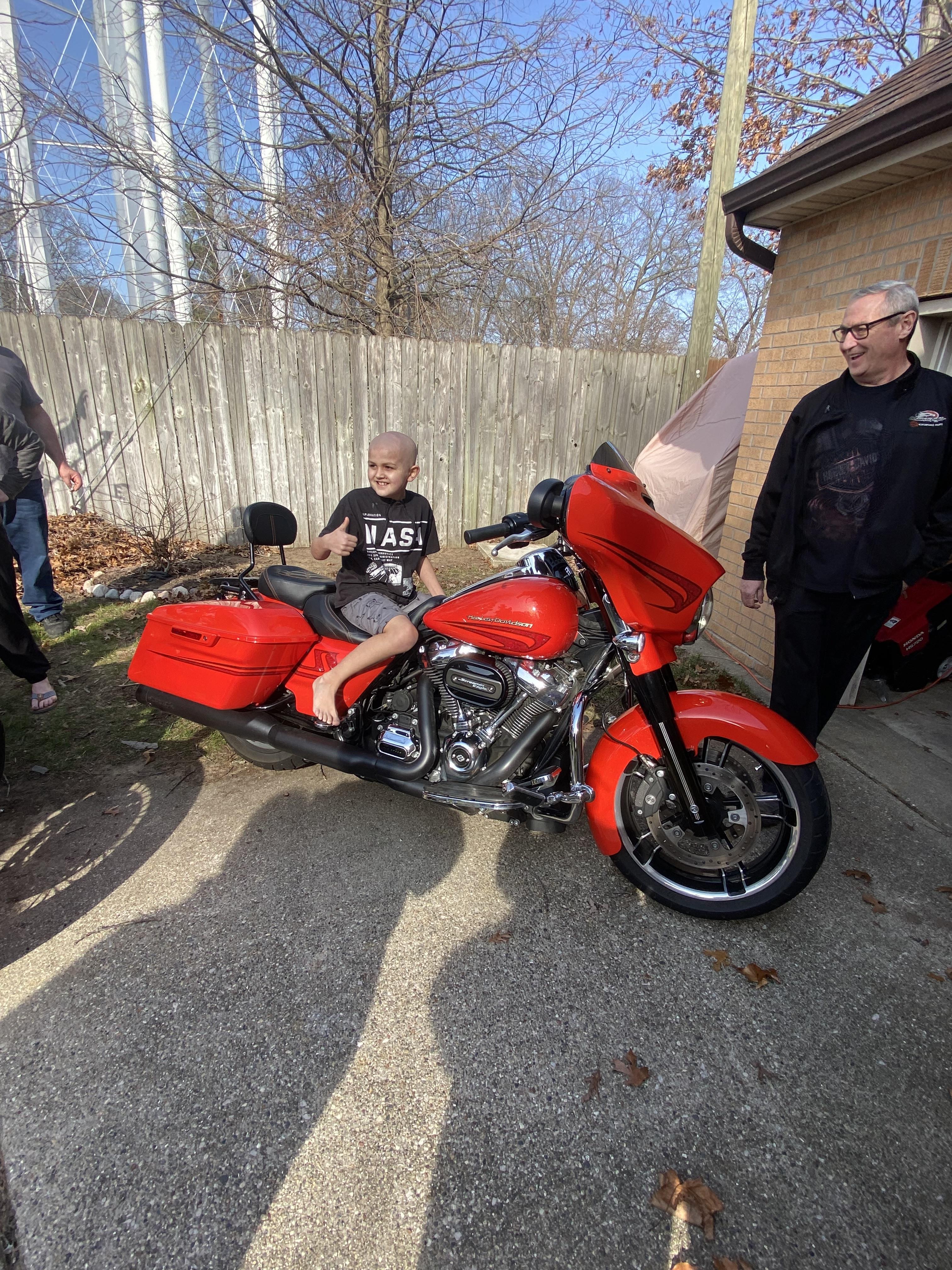 A child sits on a bright red motorcycle, surrounded by adults, in a driveway.