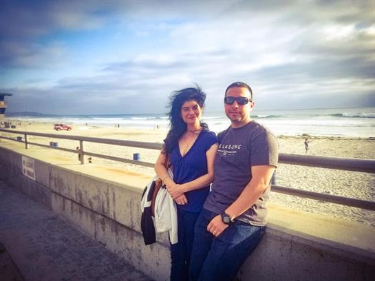 A couple poses on a boardwalk with the ocean and beach in the background, enjoying a sunny day.