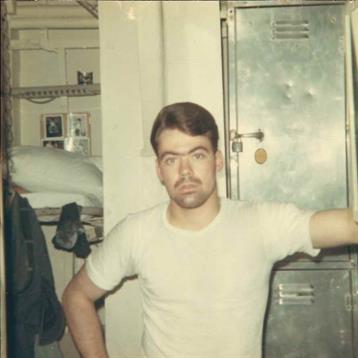 A young man stands with arms crossed in a military barracks, showcasing his confident demeanor.