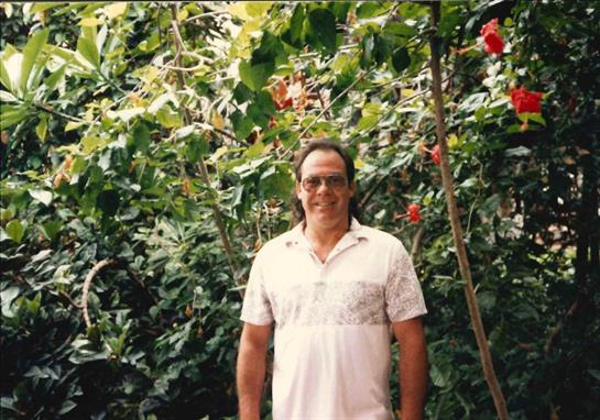 A middle-aged man poses smiling in a tropical garden filled with colorful flowers and thick foliage.