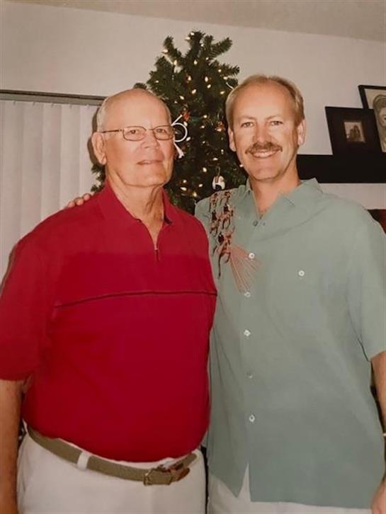Two men stand together, smiling, in front of a decorated Christmas tree.