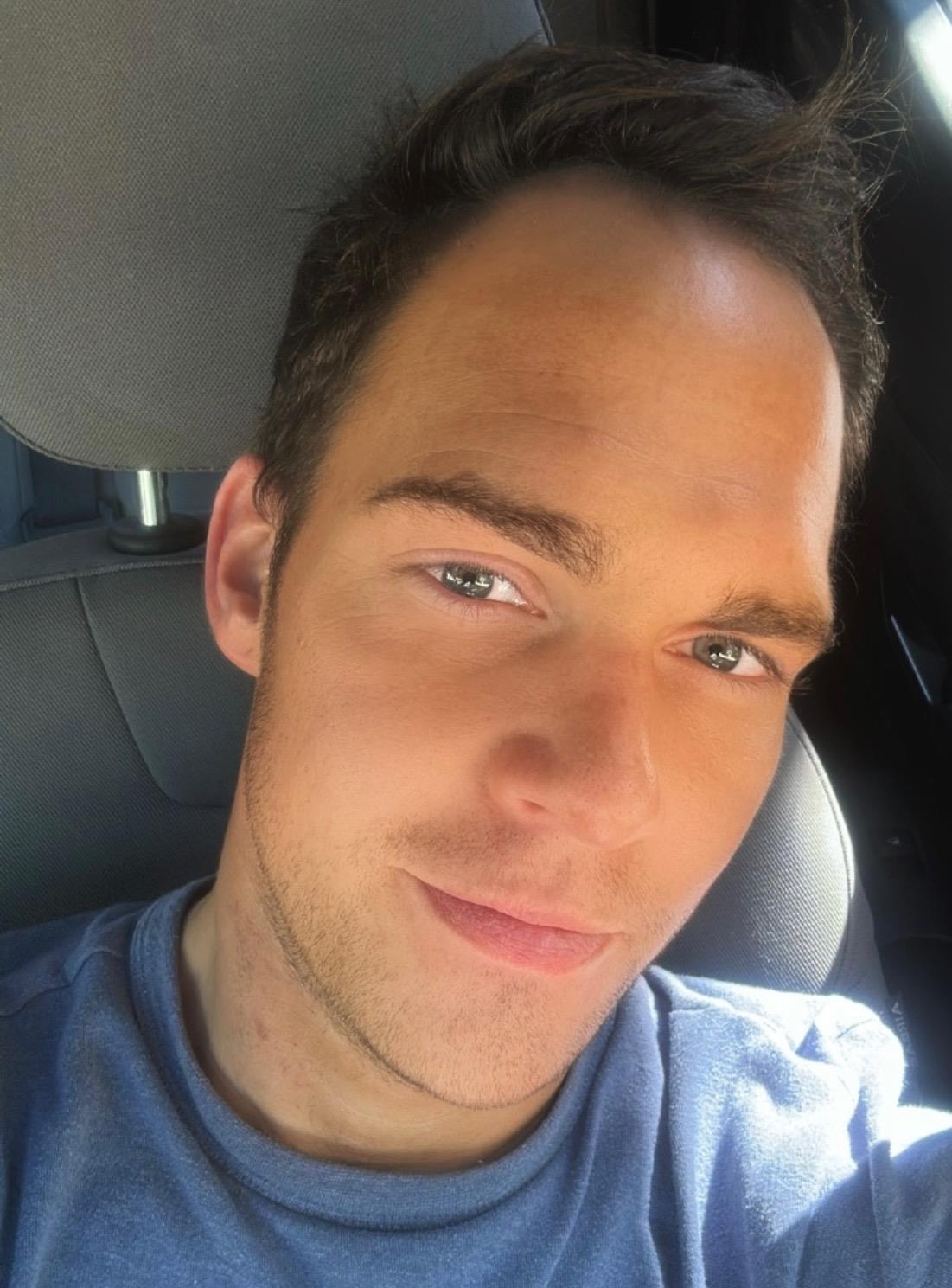 A young man captures a selfie in a car, showing a relaxed expression and casual style.
