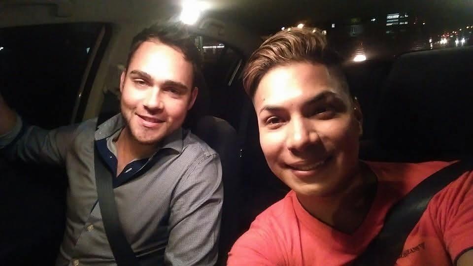 Two men smile and pose for a selfie while sitting in a car at night, illuminated by city lights.