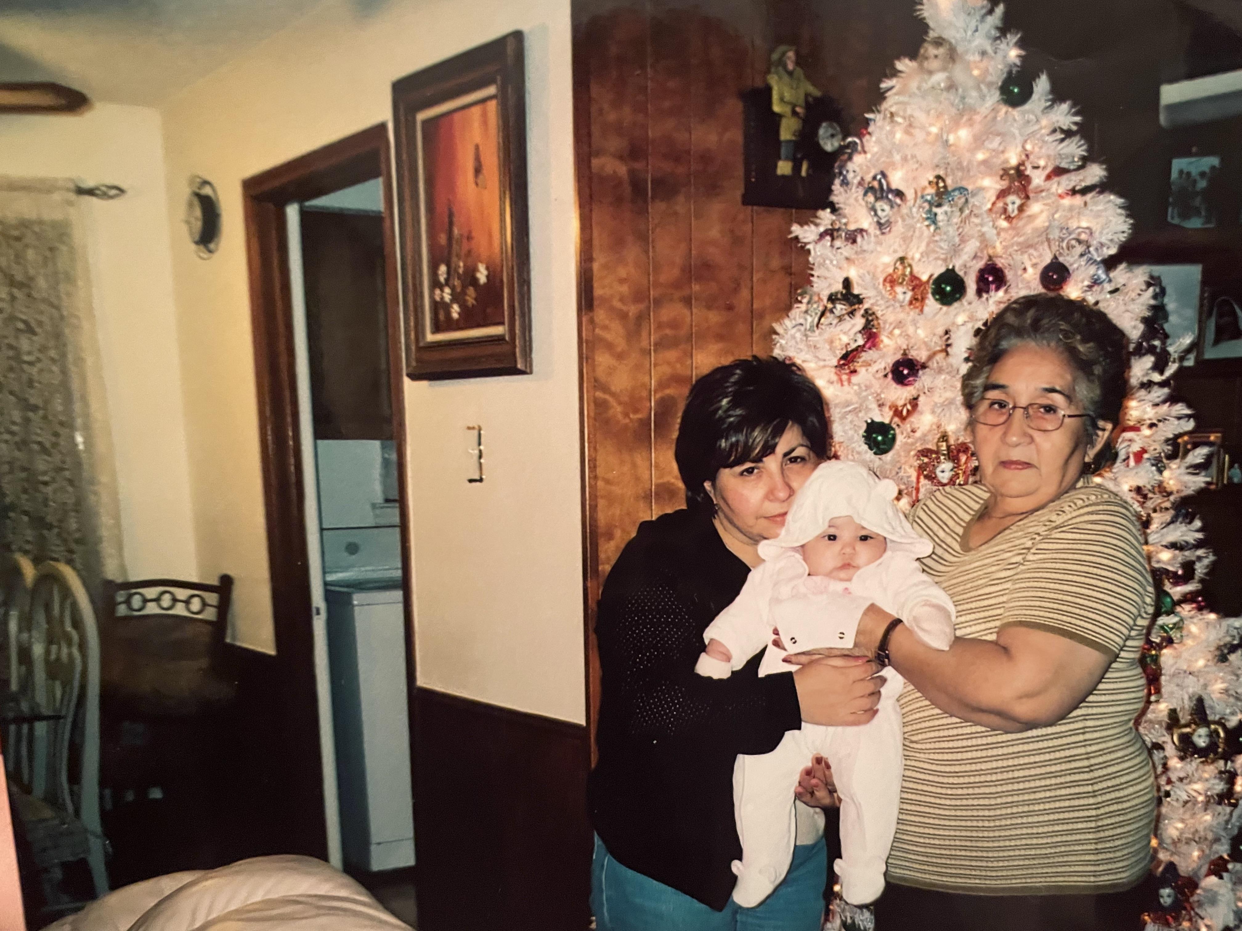 Two women hold a baby in white clothing near a bright Christmas tree in a warm home setting.