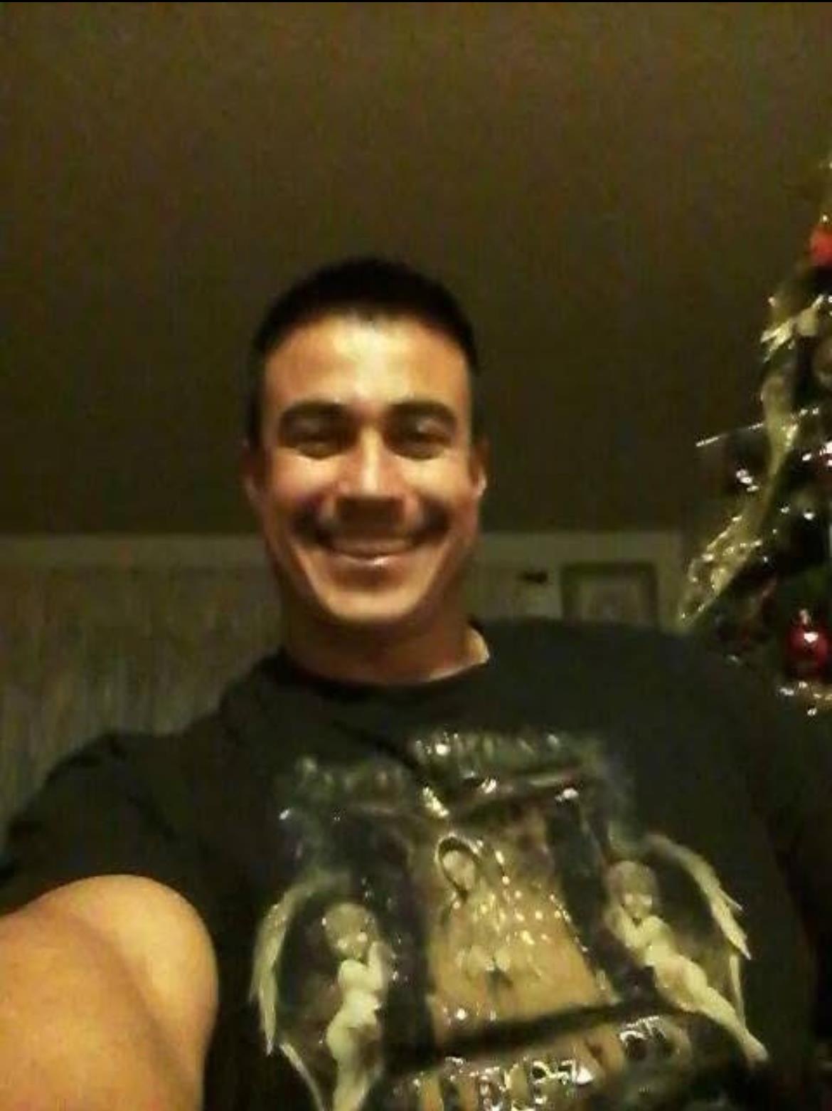 A man is joyfully posing with a Christmas tree decorated with ornaments at his home.