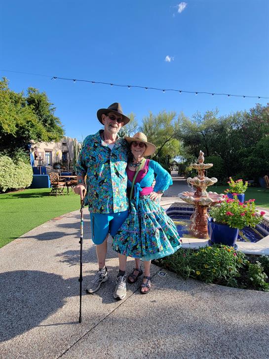 A cheerful couple stands in a colorful garden, dressed in matching outfits under the bright sun.
