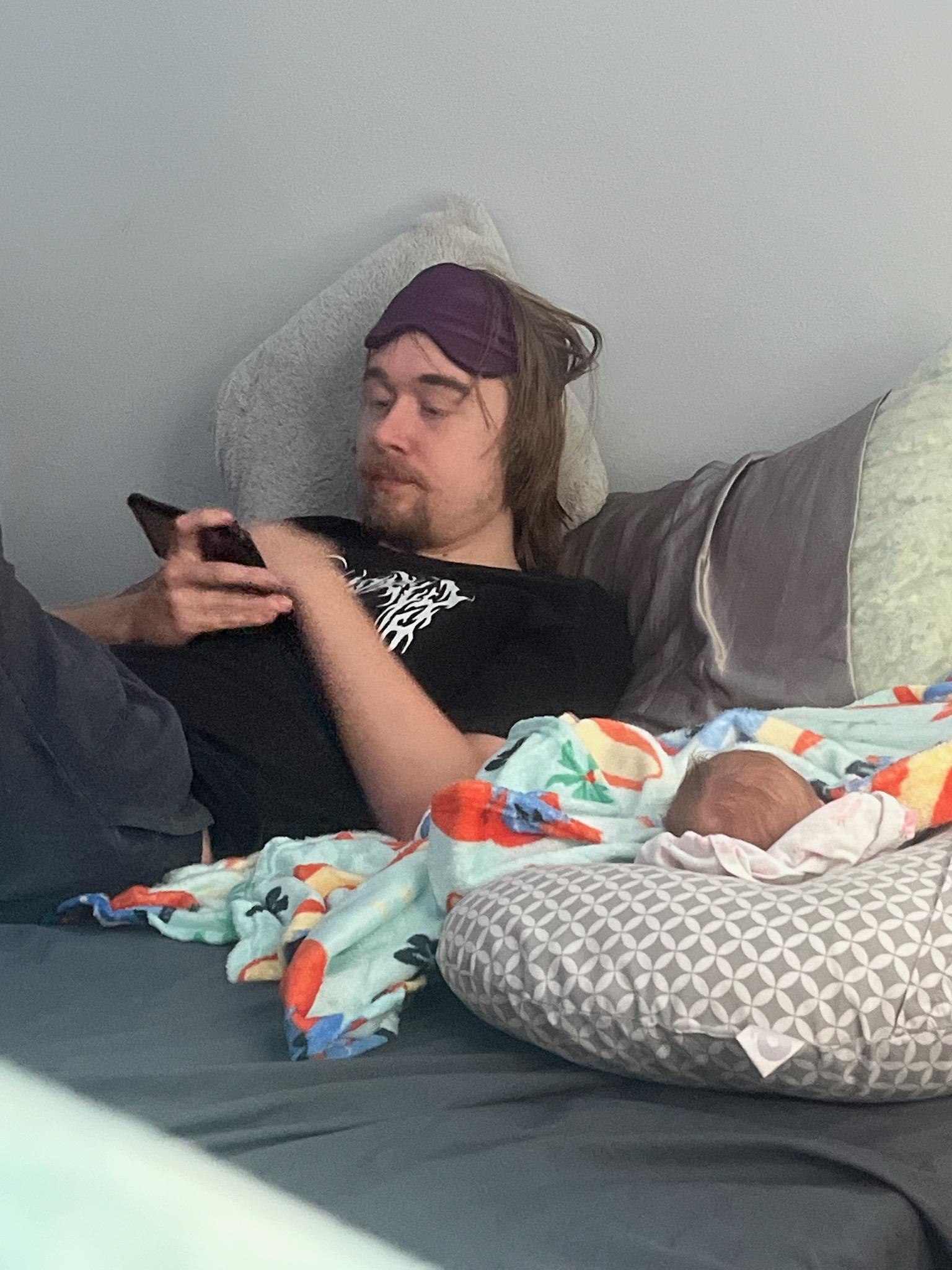 A relaxed young man uses his smartphone while lying on a bed next to a sleeping infant.