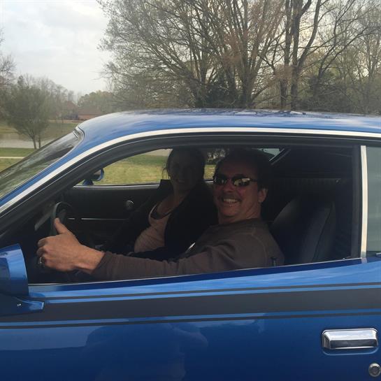 A couple smiles and poses in a blue classic car while driving through a scenic area.