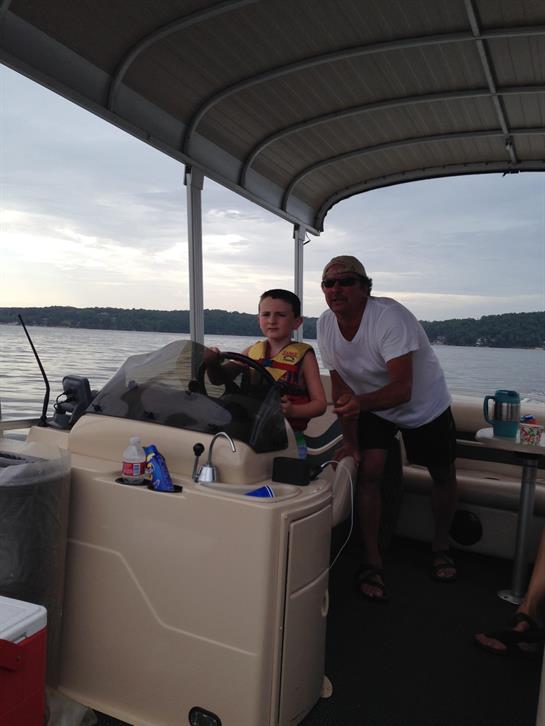 A young boy is happily steering a boat under a cloudy sky with an adult guiding him.