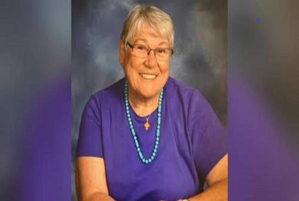 A cheerful woman with gray hair smiles warmly wearing a purple shirt and blue necklace.