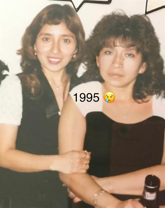 Two women stand close, smiling gently, dressed in stylish outfits from 1995 at a social gathering.