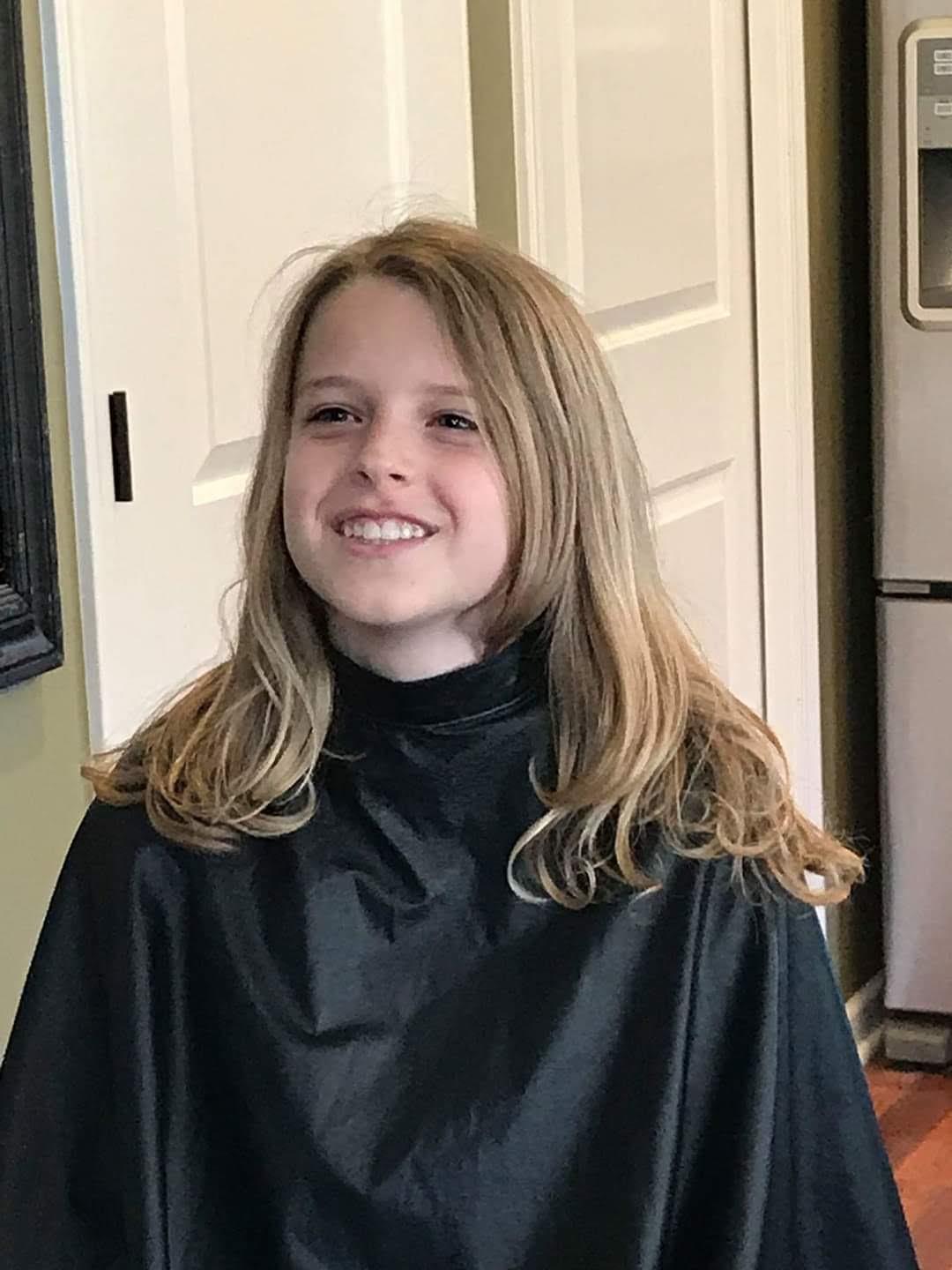 A girl with long hair beams as she sits in a salon cape, ready for her haircut.